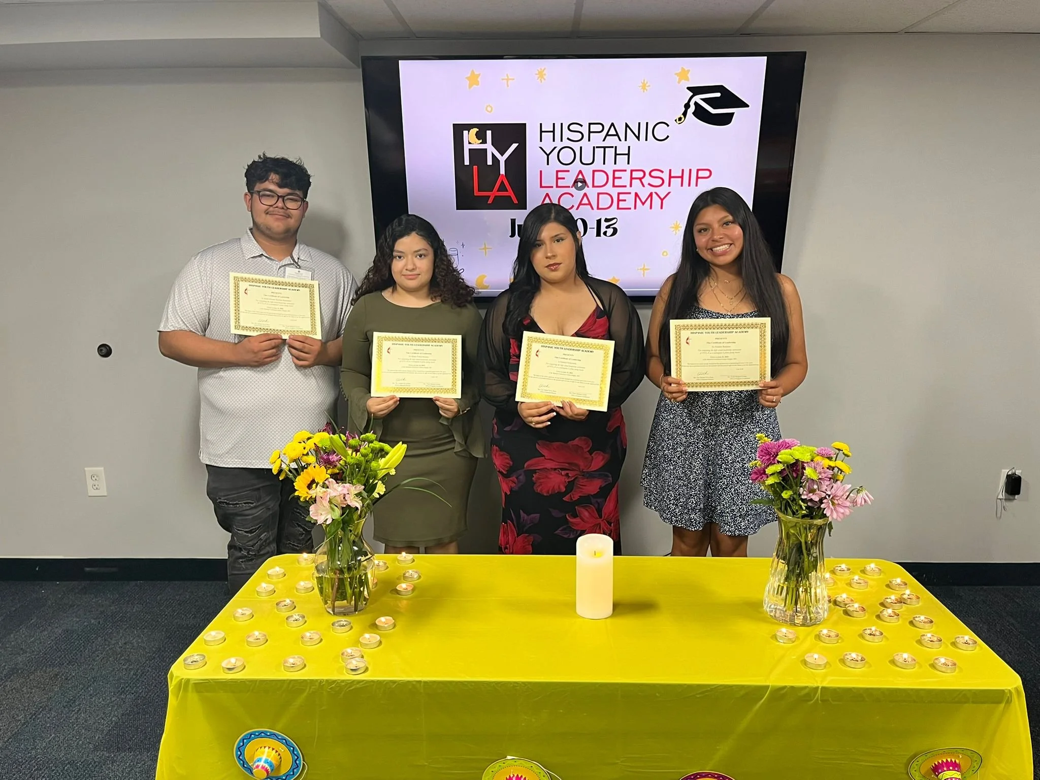 Four young adults standing behind a yellow table with flowers and candles, holding certificates, at a Hispanic Youth Leadership Academy event, with a screen behind them displaying the event's logo and name.