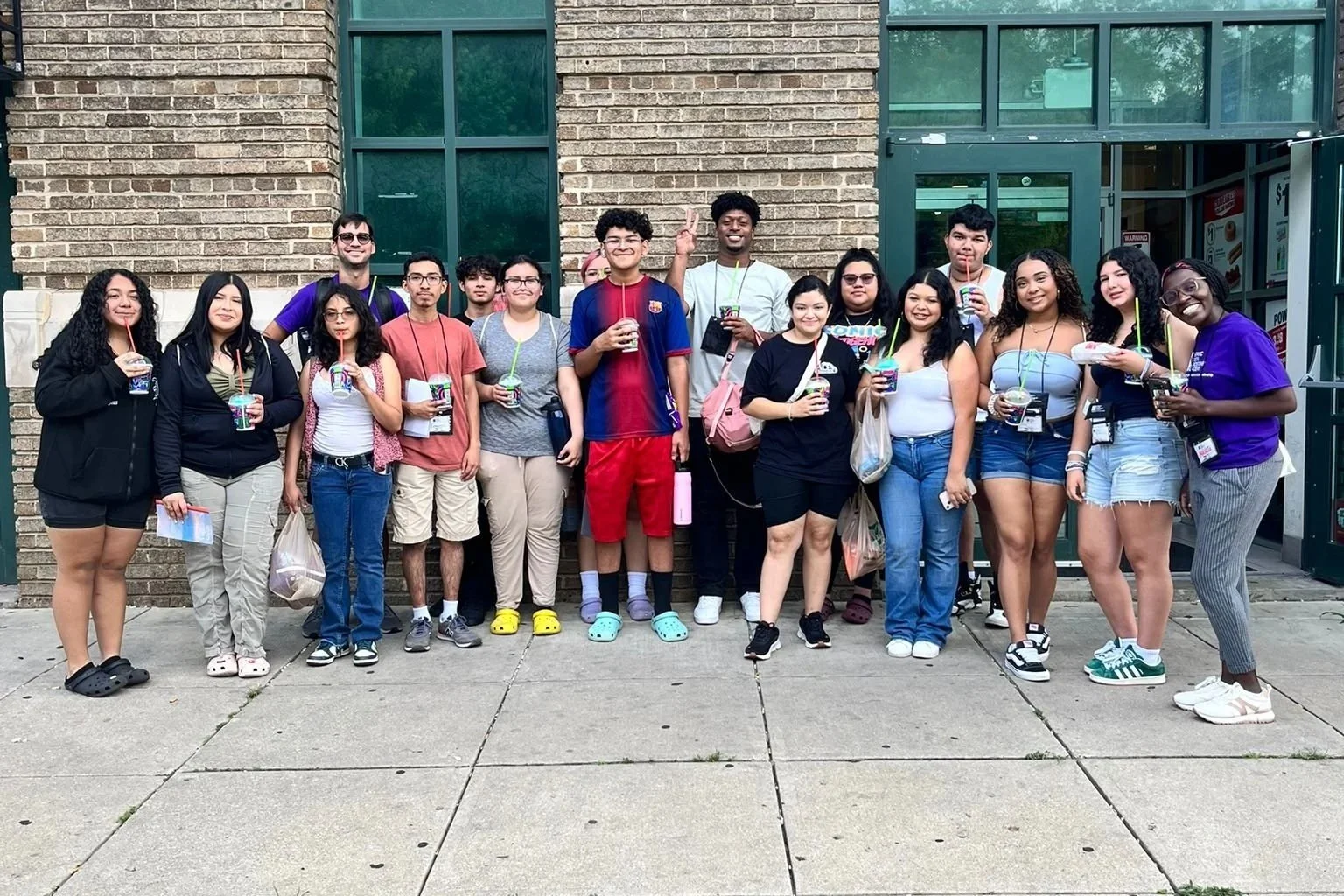 A group of around 16 young people standing outside in front of a brick building, holding drinks in cups with straws, smiling and posing for a photo.
