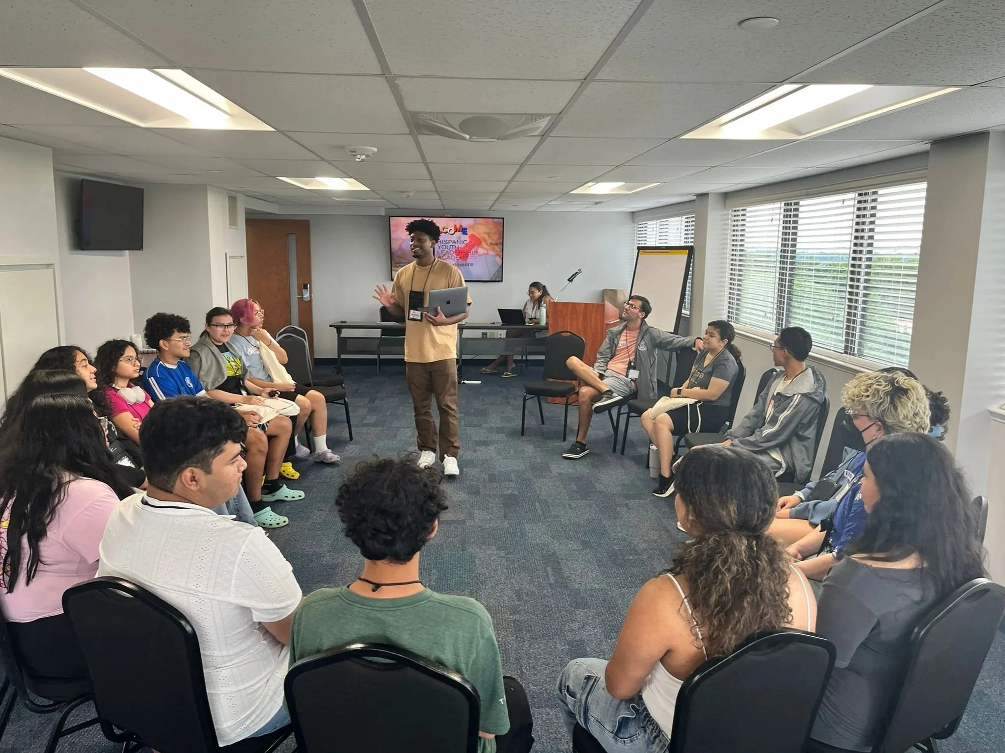 A diverse group of children and teenagers sitting in a circle in a conference room, listening to a young man standing and speaking, with a large monitor behind him showing a colorful presentation slide.