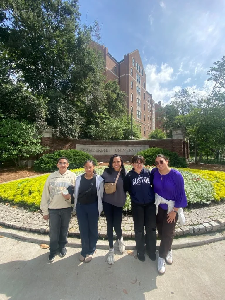 HYLA youth and facilitators pose together in front of a Vanderbilt University sign, with colorful flower beds, trees, and a brick building in the background.