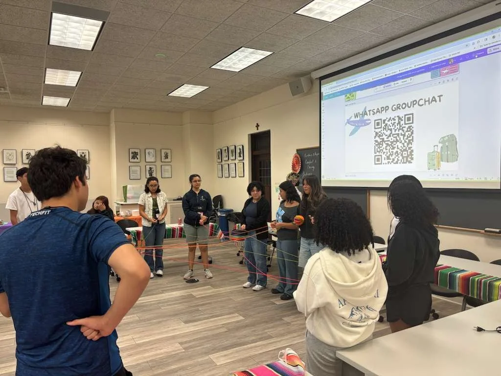 Group of students participating in an activity with a red string in a classroom, with a large screen showing a WhatsApp group chat QR code and luggage icons.