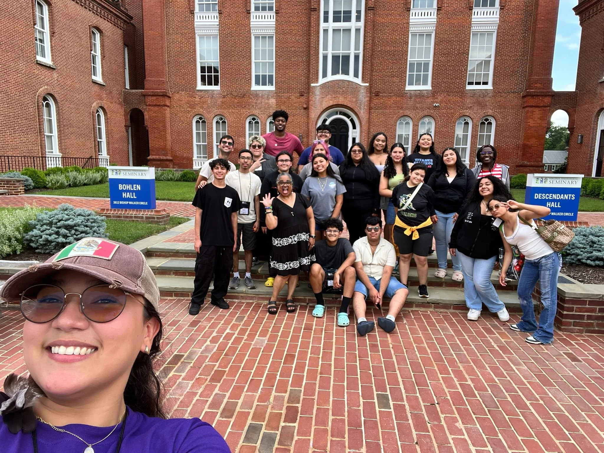Group of young people posing for a photo outside Bohen Hall at Southeastern Seminary, with a woman taking a selfie in the foreground. The group is standing on brick stairs in front of a large red brick building with signs indicating Bohen Hall and De