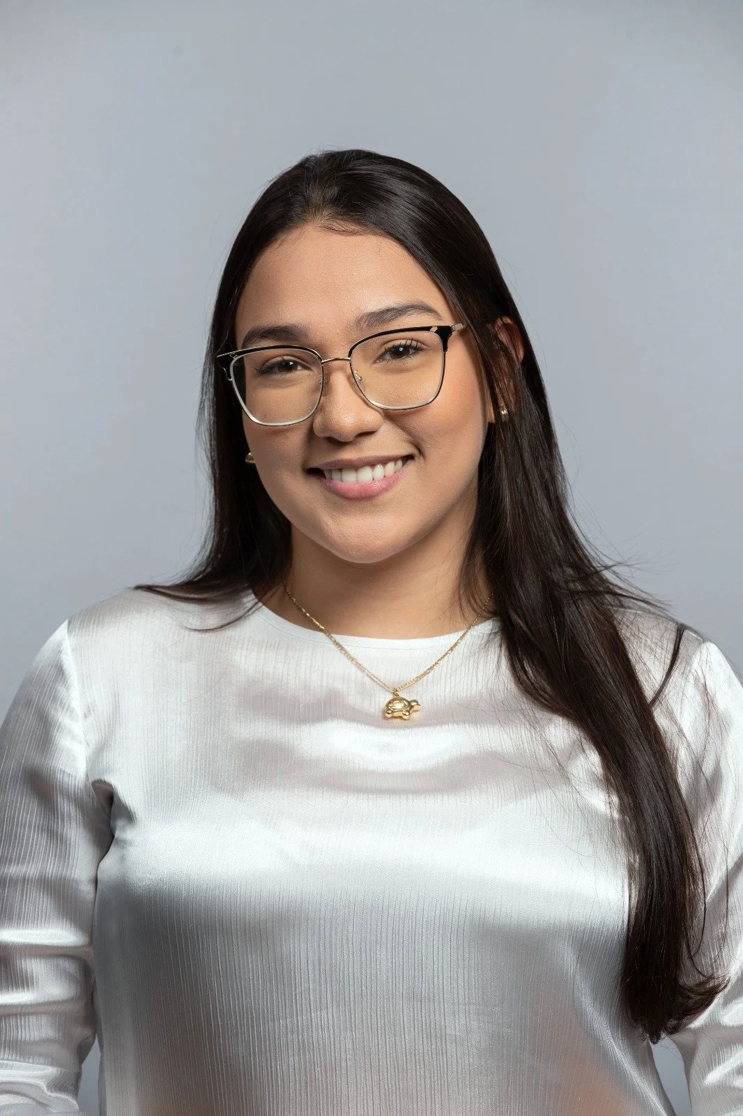 A young woman with long dark hair, glasses, and a warm smile, wearing a white satin top and a gold necklace with a car pendant, standing against a neutral gray background.