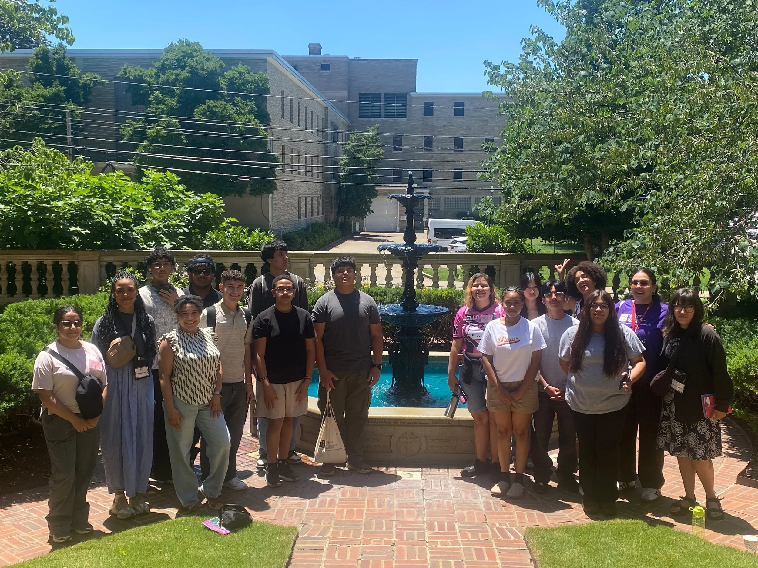 HYLA youth and facilitators pose for a photo around a tiered fountain in a sunny university courtyard, surrounded by green trees, shrubs, and campus buildings.