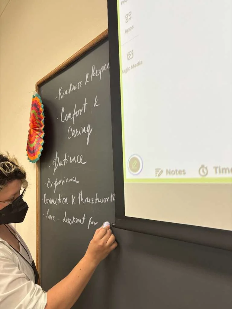 A woman writing on a blackboard with a piece of chalk, with a black mask covering her mouth and nose, and a black and white shirt.