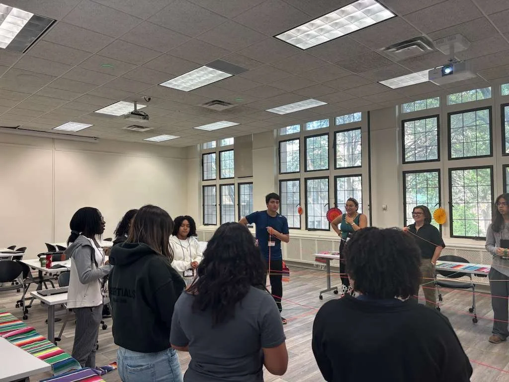 A group of people standing in a circle in a brightly lit room with large windows, participating in a team-building activity involving color-coded ropes.