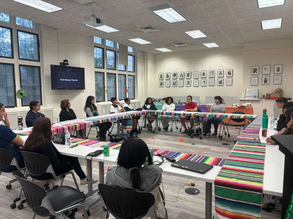 A group of people sitting in a U-shaped arrangement of tables in a conference room with large windows, a TV on the wall, and framed artwork. Some tables are covered with colorful striped tablecloths.