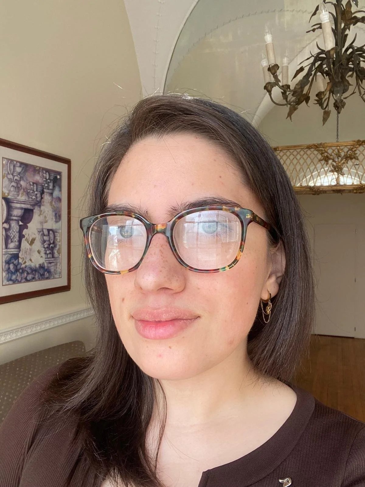 Close-up of a young woman with brown hair, glasses with a tortoiseshell pattern, and earrings, standing indoors with a framed picture and chandelier in the background.