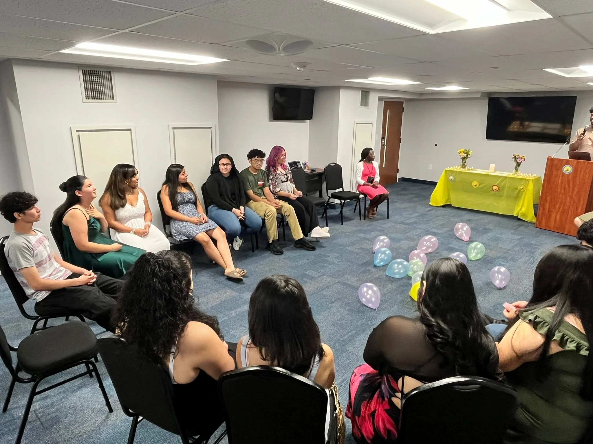 People sitting in chairs watching a woman speaking at a podium in a decorated room with pastel-colored balloons on the floor.
