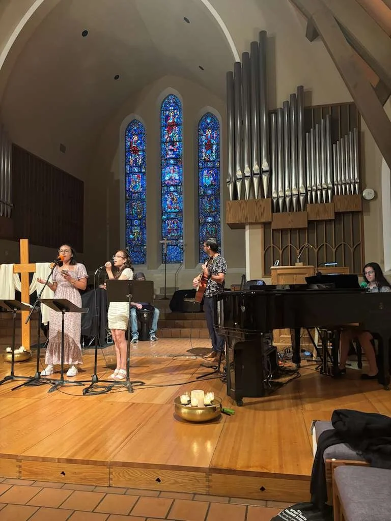 A group of people performing music on a stage inside a church, with stained glass windows and church organ pipes in the background.