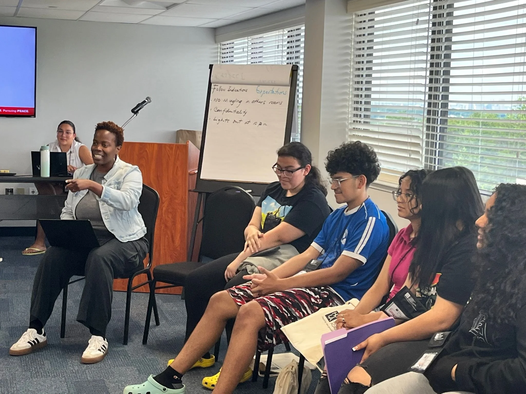 Group of five people attending a presentation in a conference room, with a woman in the front speaking and others listening.