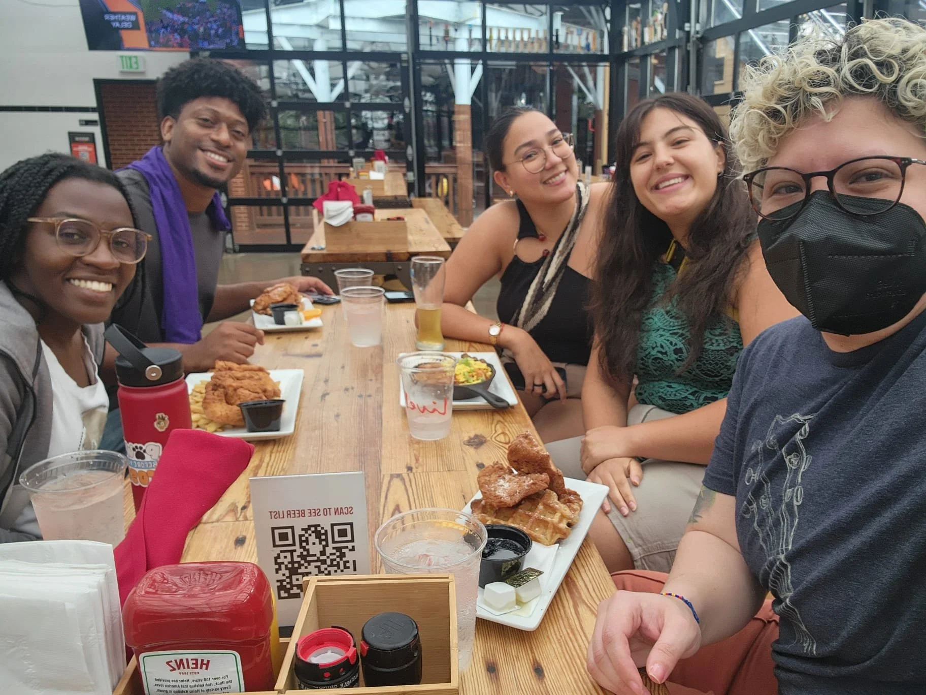 Group of six friends sitting at a wooden table in a restaurant, enjoying food and drinks. Someone is wearing a face mask, and there are plates of fried chicken, waffles, and various beverages on the table.