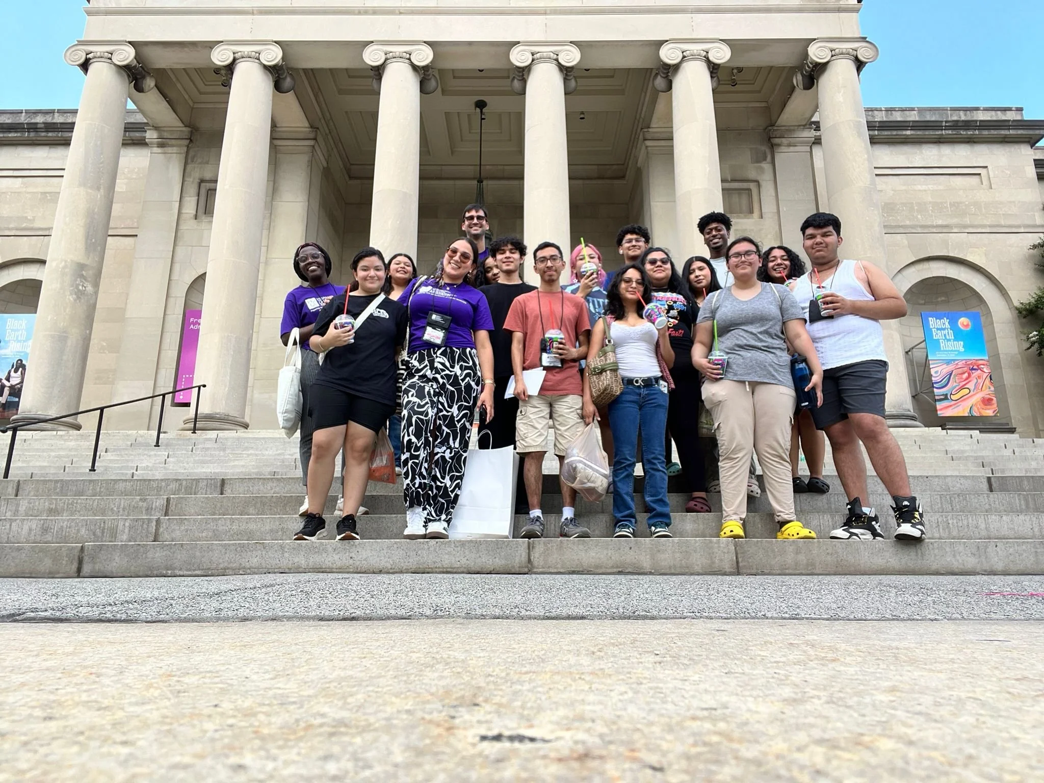 A group of people standing on the steps of a building with tall columns, holding drinks, and posing for a photo.