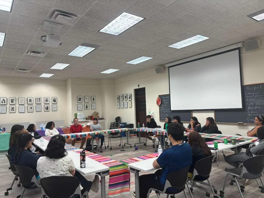 A group of people seated around U-shaped tables in a classroom or meeting room, with a large presentation screen and chalkboard at the front.