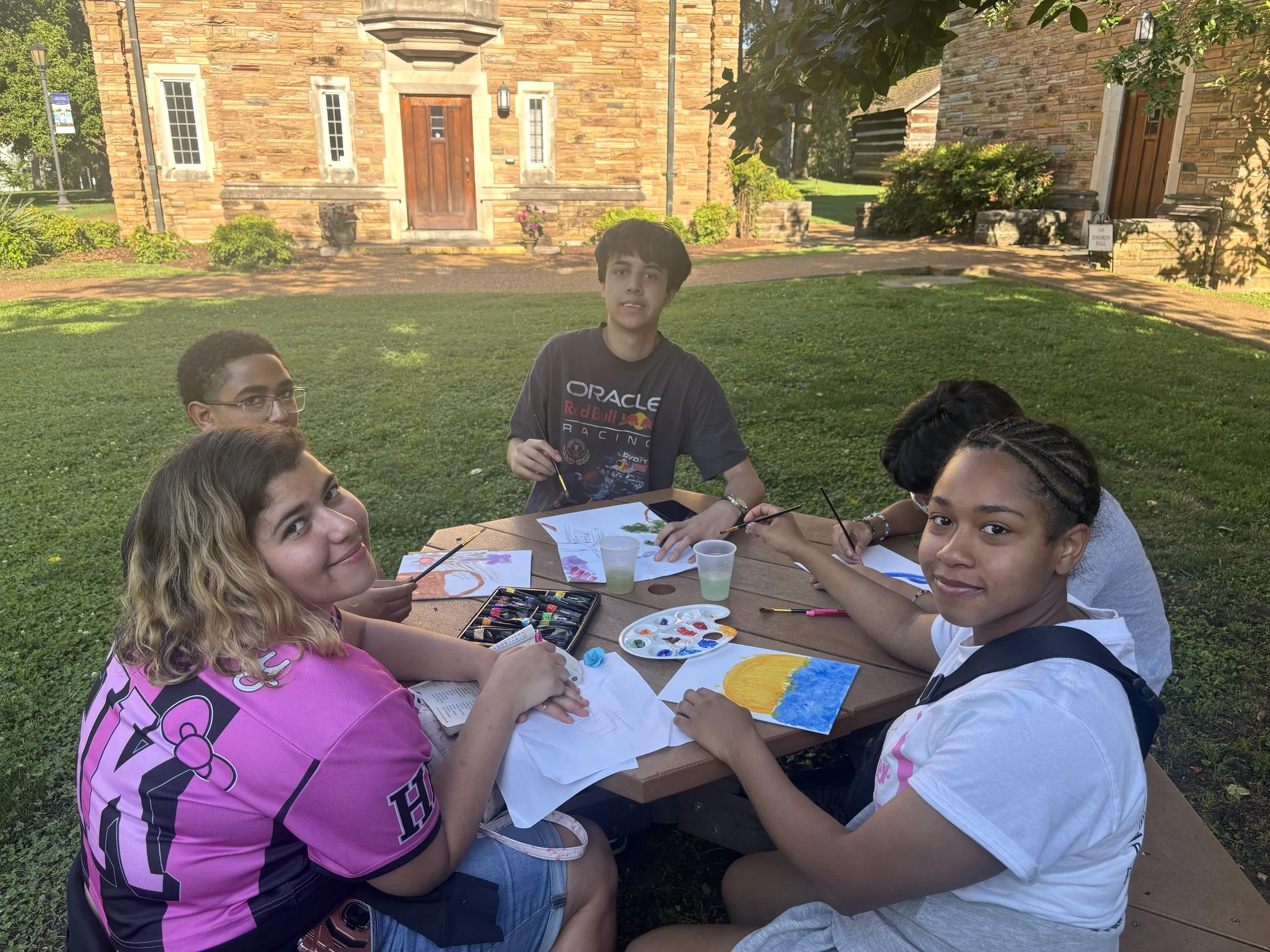 HYLA youth sit around a picnic table outdoors, painting pictures with watercolors and art supplies in front of a stone building.