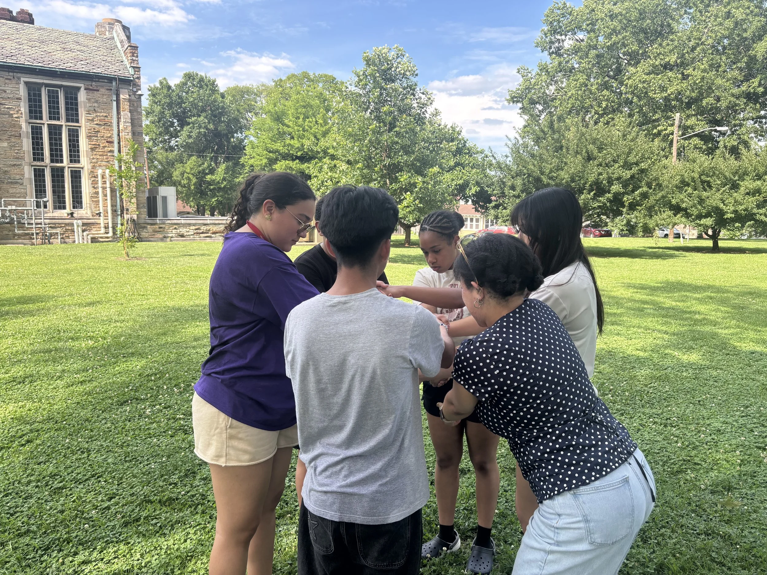 HYLA youth and staff stand in a circle on a grassy campus lawn, leaning inward and joining their hands together, with trees and a stone university building in the background.