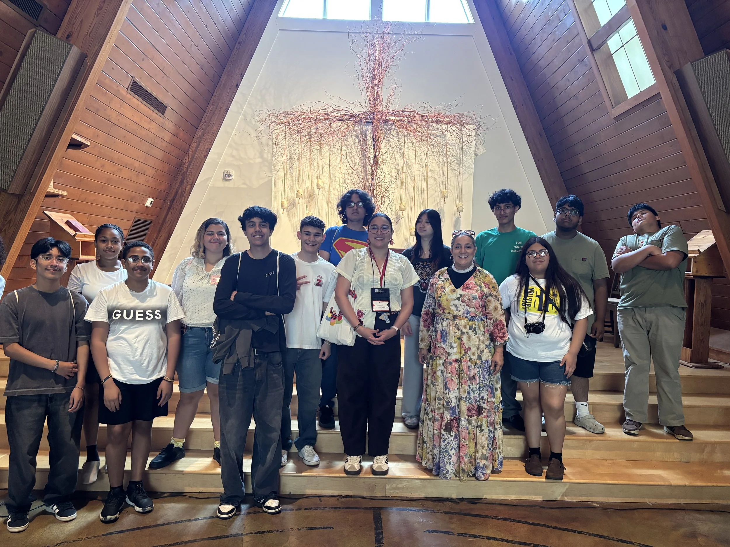 HYLA youth and staff pose for a photo on wooden steps inside the Upperroom Chapel in Nashville, TN, beneath a large cross-shaped artwork and tall windows in a high, wood-paneled ceiling.