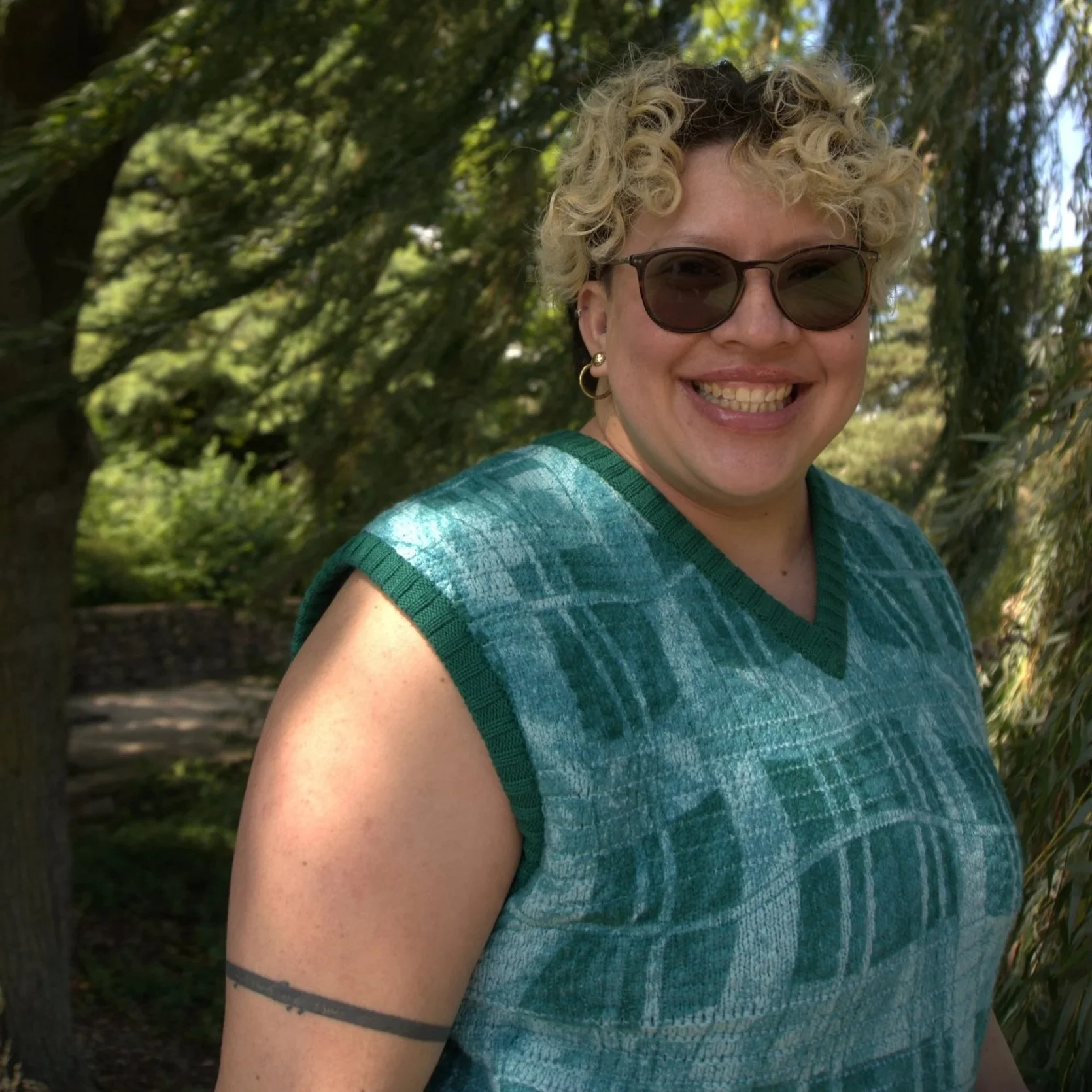 A smiling woman with curly blonde hair wearing sunglasses, a blue patterned sleeveless shirt, and earrings, standing outdoors in a lush green park with trees in the background.
