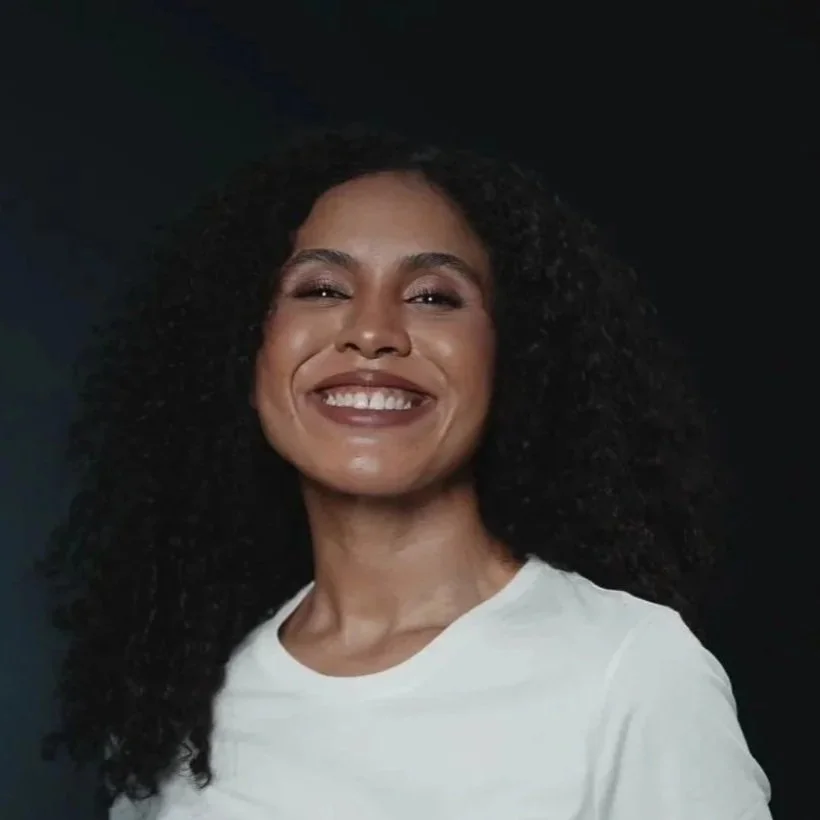 A woman with curly black hair smiling against a black background, wearing a white shirt.