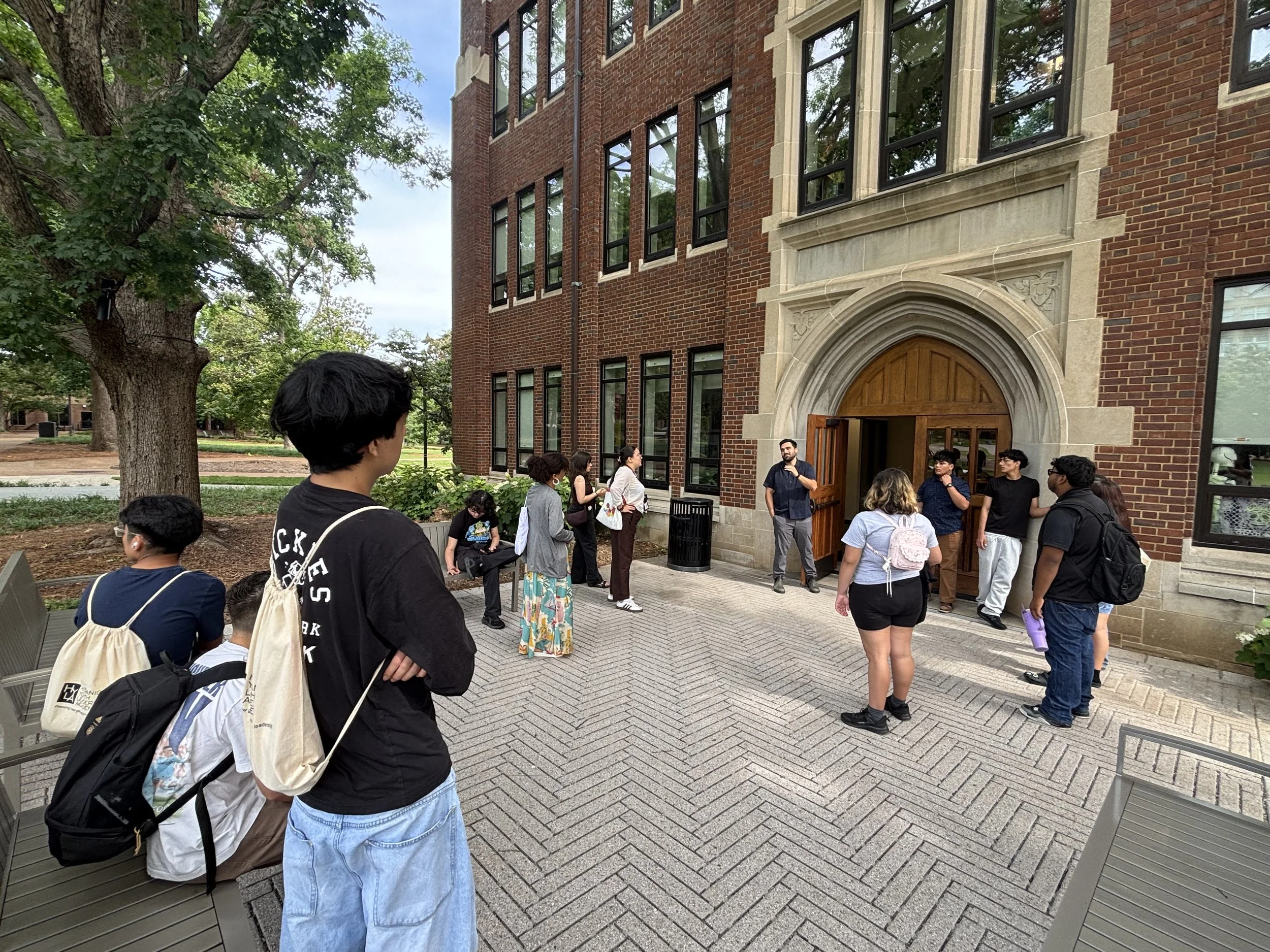 HYLA youth gather outside a red-brick university building with an arched stone entrance, listening to a guide speak near the open doorway.