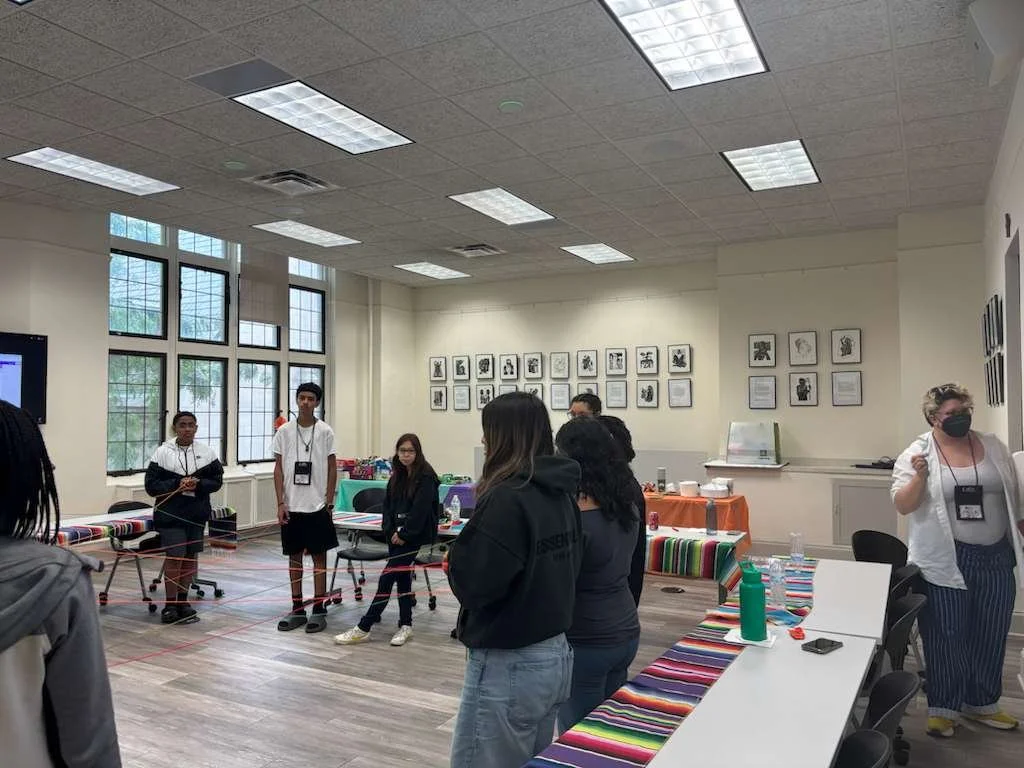 A group of people in a room engaging in a team-building activity involving a web of red yarn stretched between chairs.