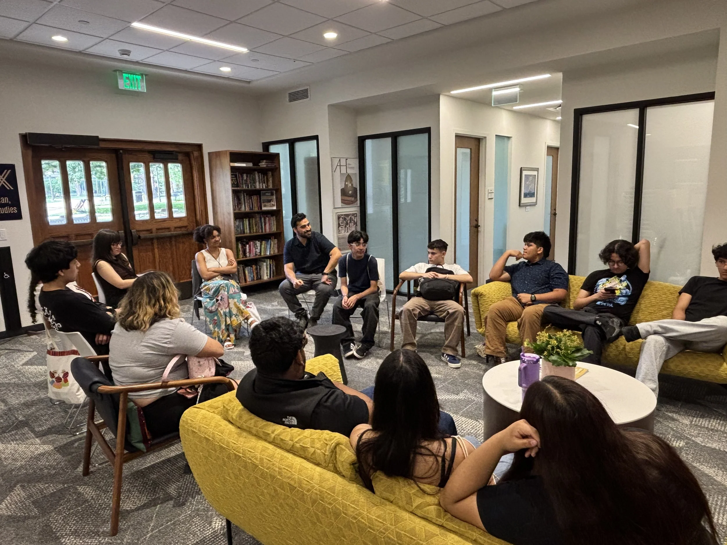 HYLA youth and facilitators sit in a circle in a campus lounge with yellow couches and chairs, in a group discussion with Dr. Jesus Ruiz of Vanderbilt University.
