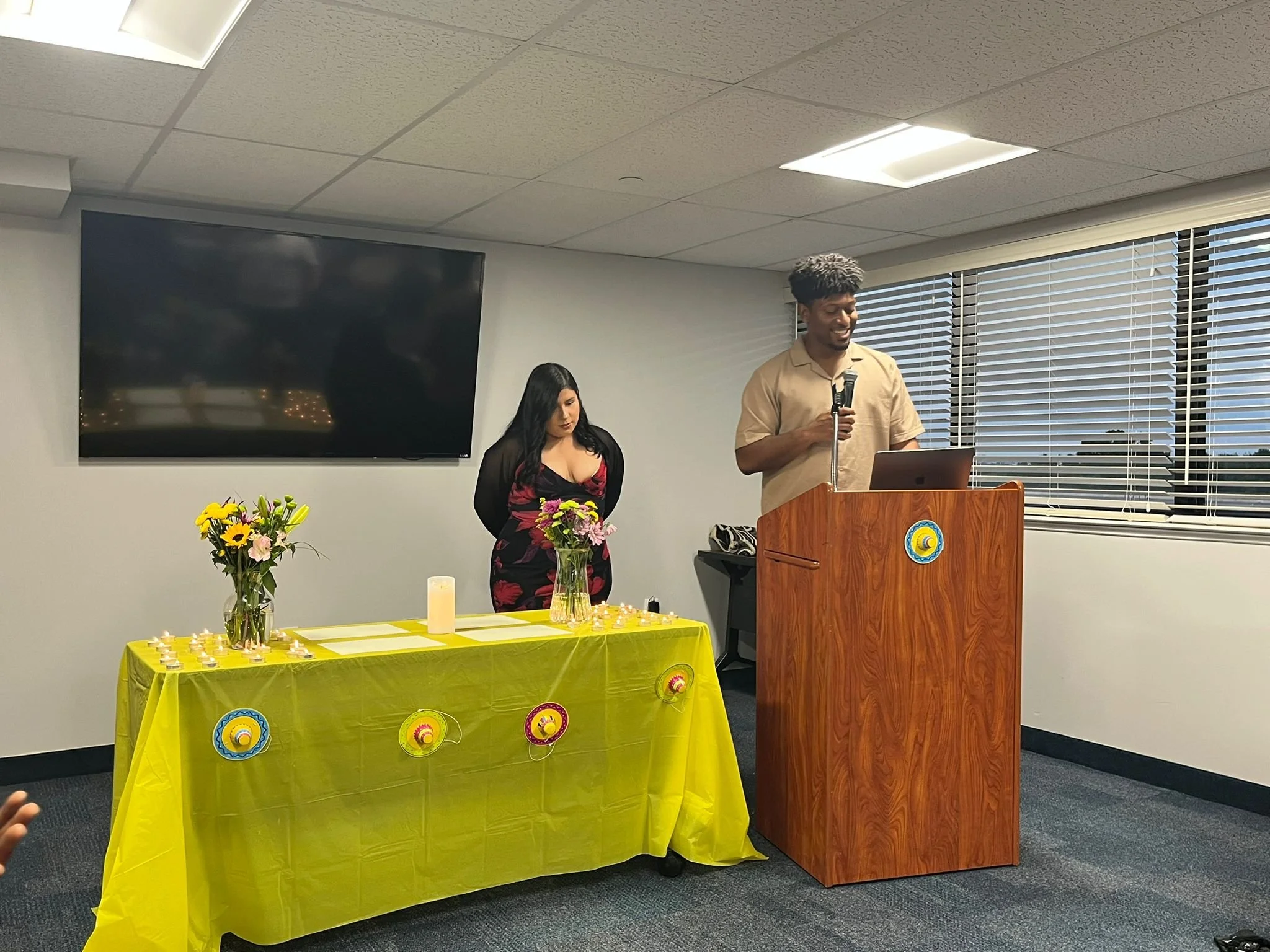 A young man standing at a wooden podium, holding a microphone and smiling, with a laptop in front of him. Next to him, a woman stands behind a yellow table decorated with flowers and colorful paper lanterns, with her hands clasped. The room has a lar
