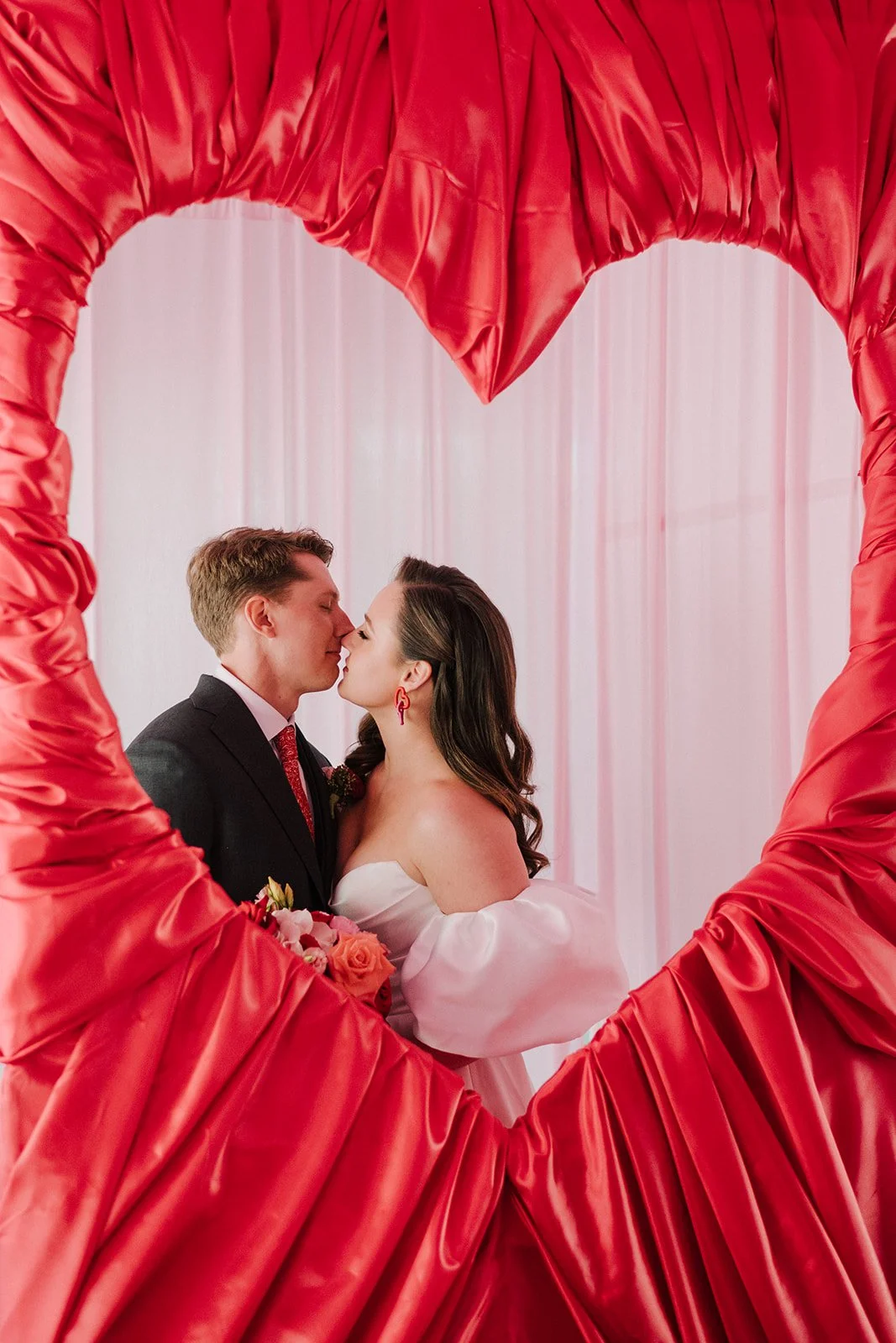 A couple dressed in wedding attire sharing a kiss inside a red heart-shaped decoration.