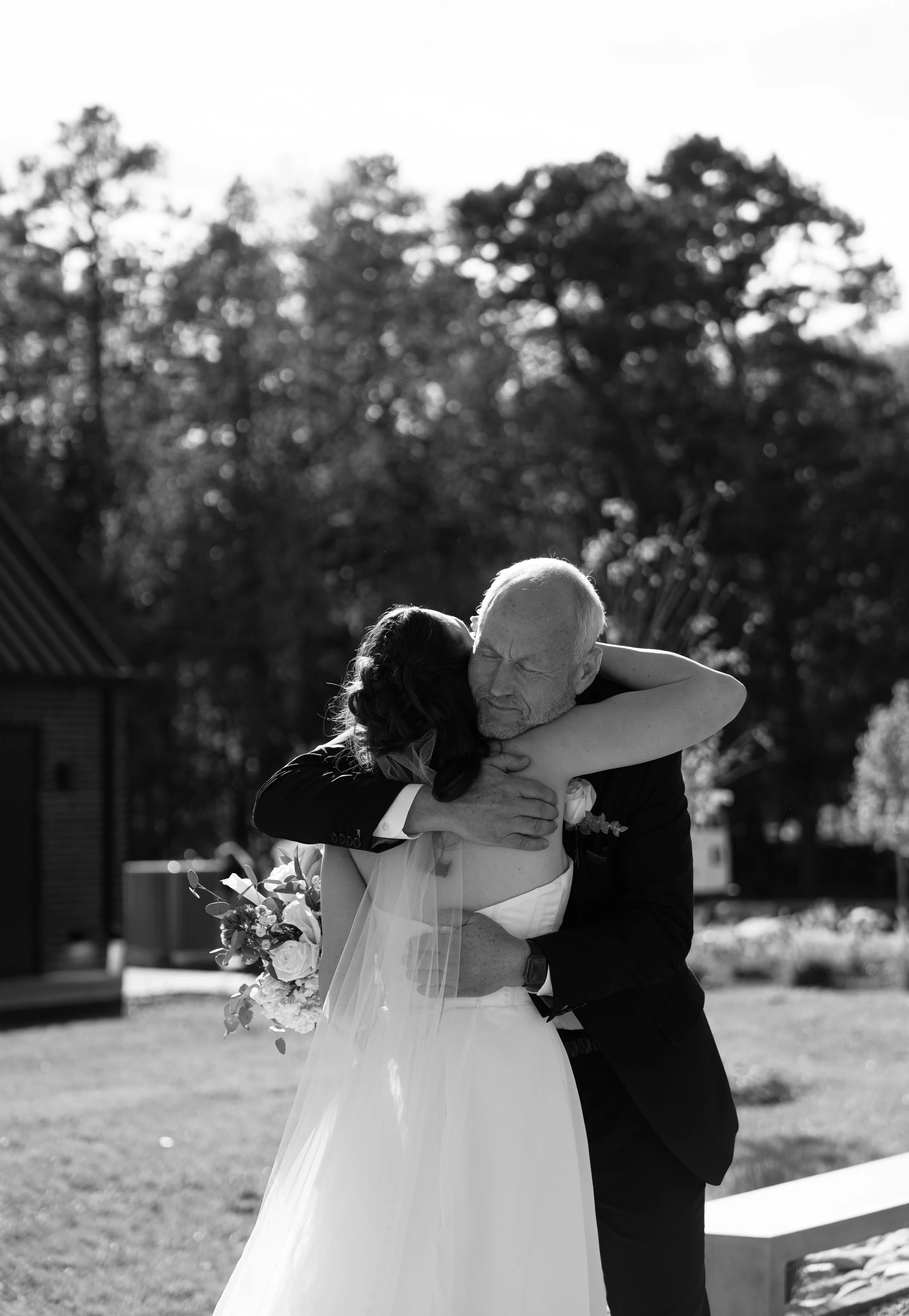 An elderly man and a woman, dressed as a bride, embrace tightly outdoors during a wedding ceremony, with trees in the background.