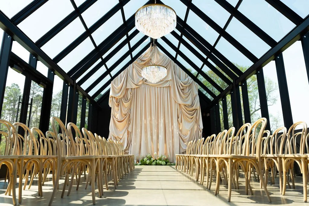 Elegant indoor wedding ceremony setup with rows of wooden chairs and a large, draped fabric backdrop under a glass ceiling with chandeliers.