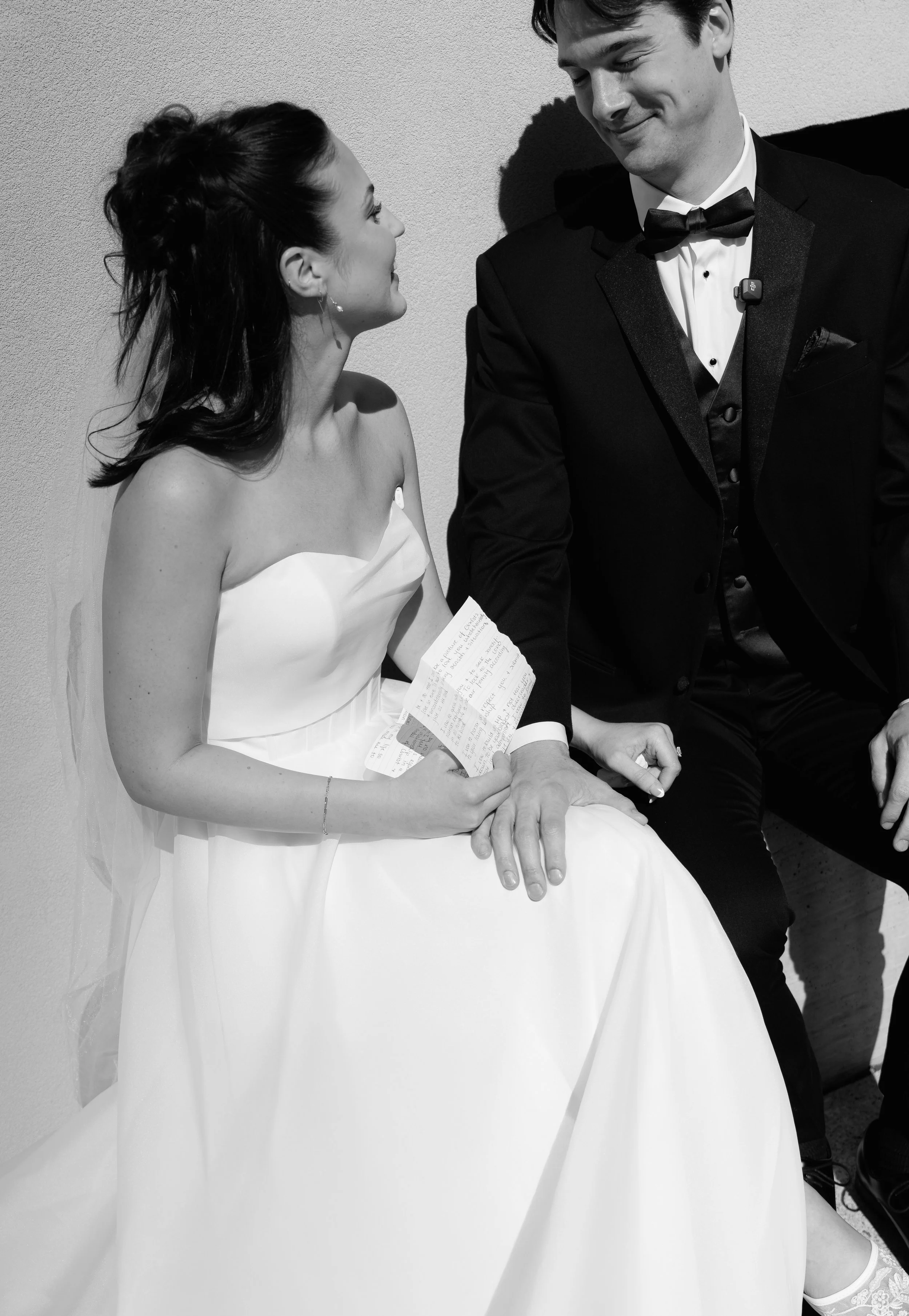 A bride and groom sitting together, holding hands, dressed in wedding attire. The bride is wearing a strapless white wedding dress, and the groom is in a tuxedo with a bow tie. The bride is holding a handwritten note, and they are looking at each oth