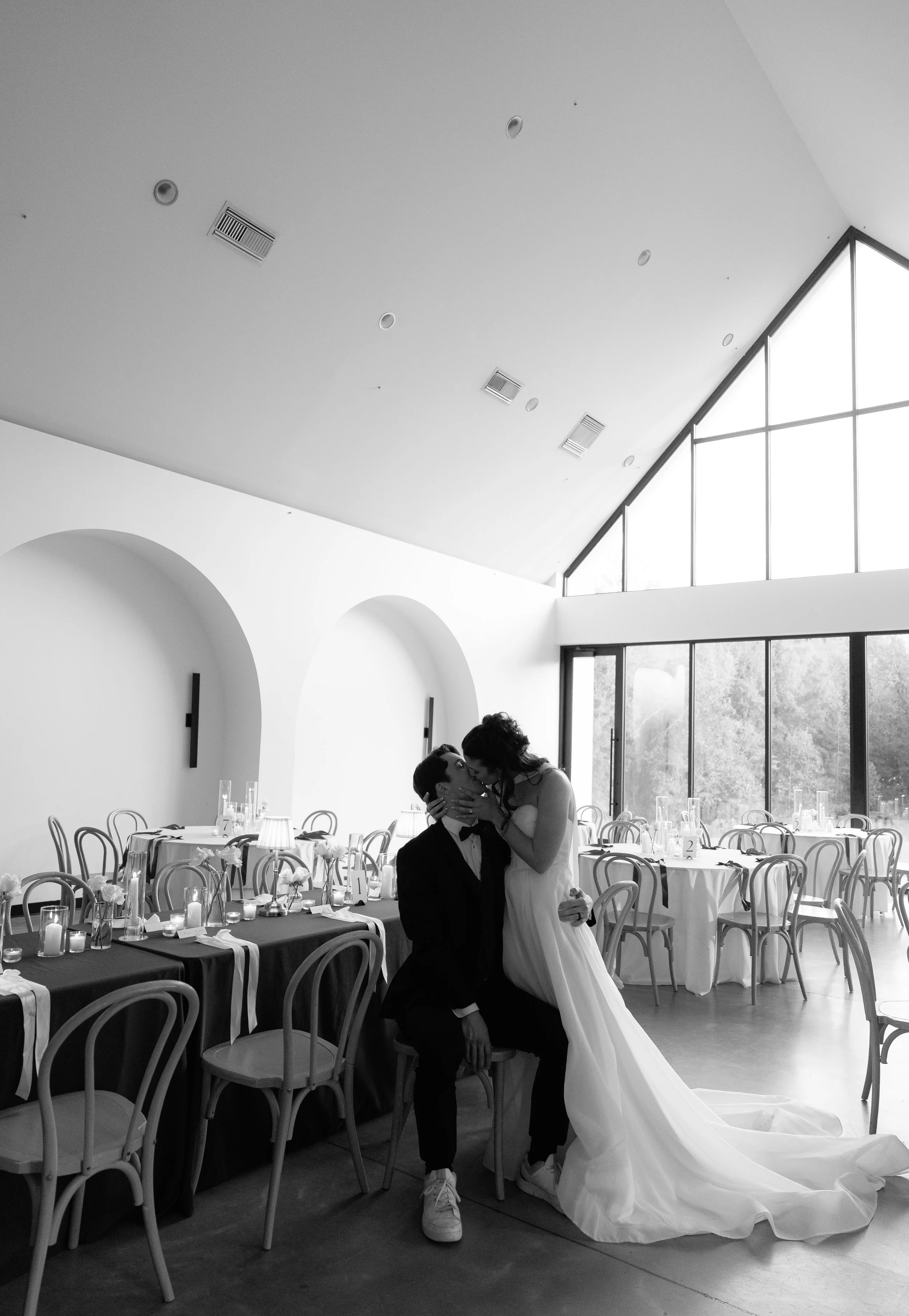 A bride and groom in wedding attire sharing a kiss in a modern, elegant reception hall with large windows and decorated tables.