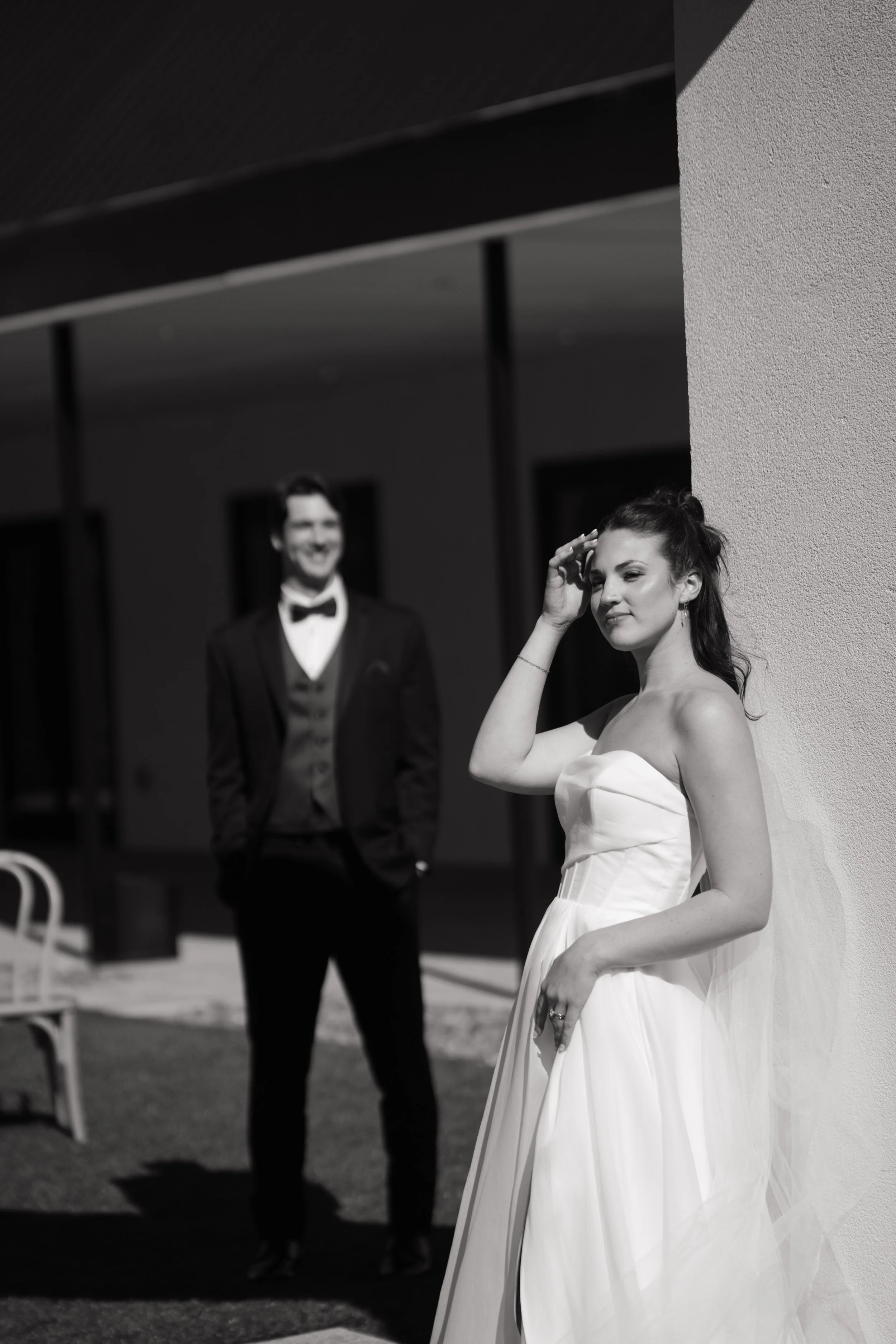 Black and white photo of a woman in a strapless wedding dress posing in front of a wall, with a man in a tuxedo smiling in the background.