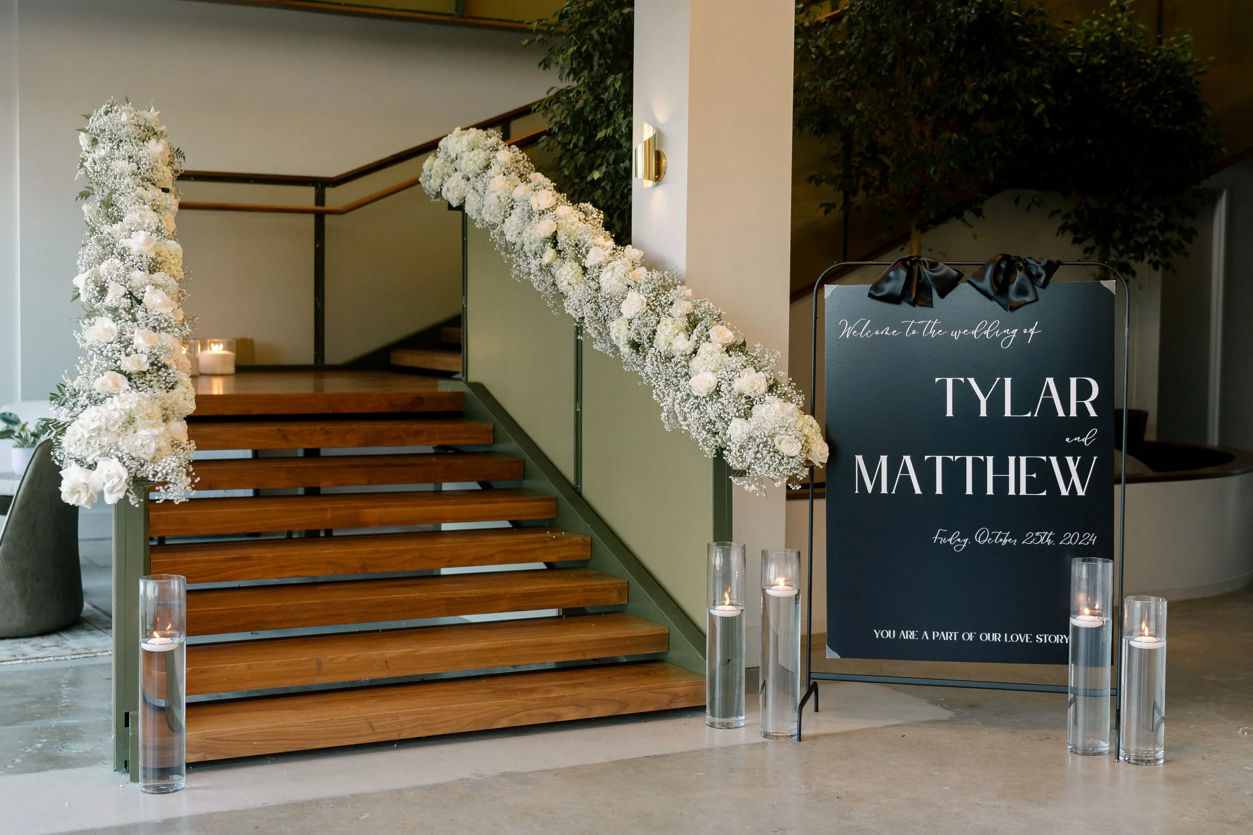 Decorated staircase for a wedding with a floral arrangement of white flowers and baby's breath on the railing, candles in glass holders at the base, and a black sign with white text welcoming guests to the wedding of Tylar and Matthew on October 25th