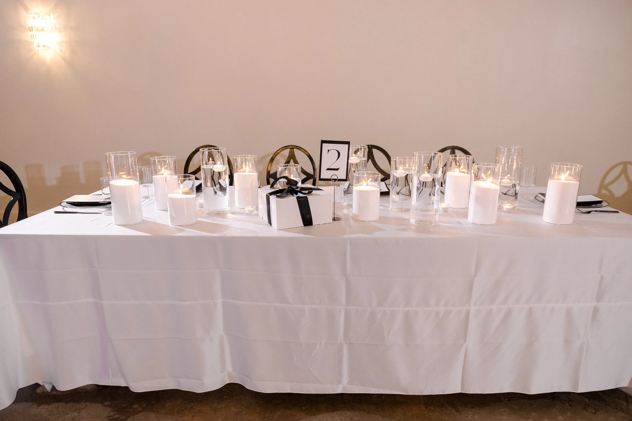 Table decorated with lit candles in glass holders, a gift box with a black ribbon, a table number card with the number 2, and place settings, set against a plain wall.