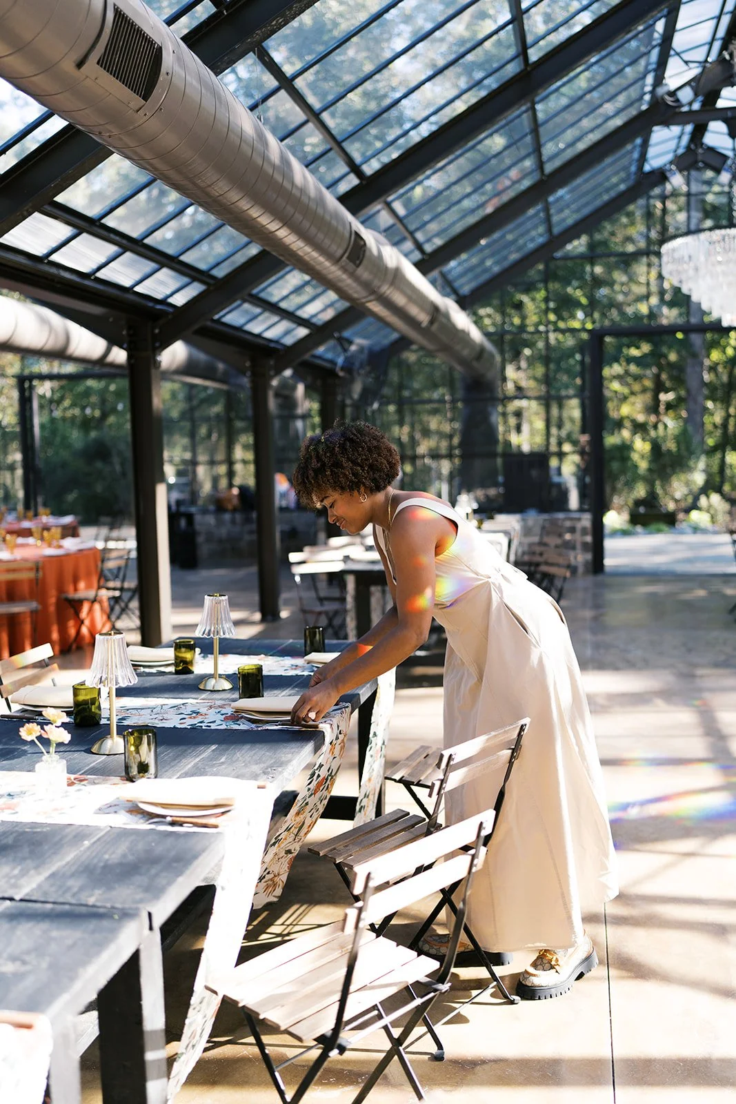 A woman in a white dress is setting a table in a bright, glass-enclosed restaurant with sunlight streaming in.