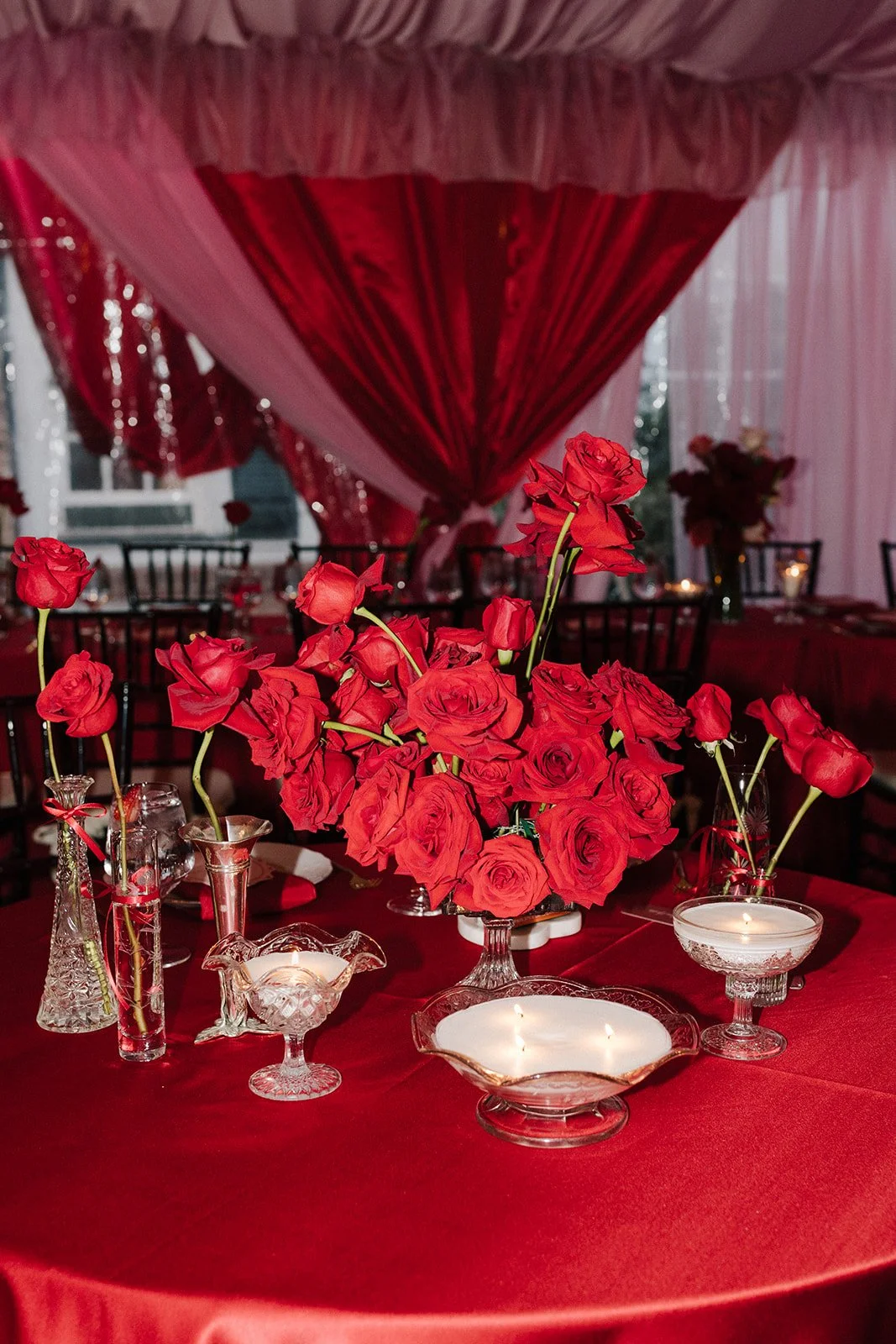 A table decorated with a large bouquet of red roses in the center, surrounded by small glass vases with single red roses, lit candles, and a red satin tablecloth. The background features red and pink draped curtains and chairs.