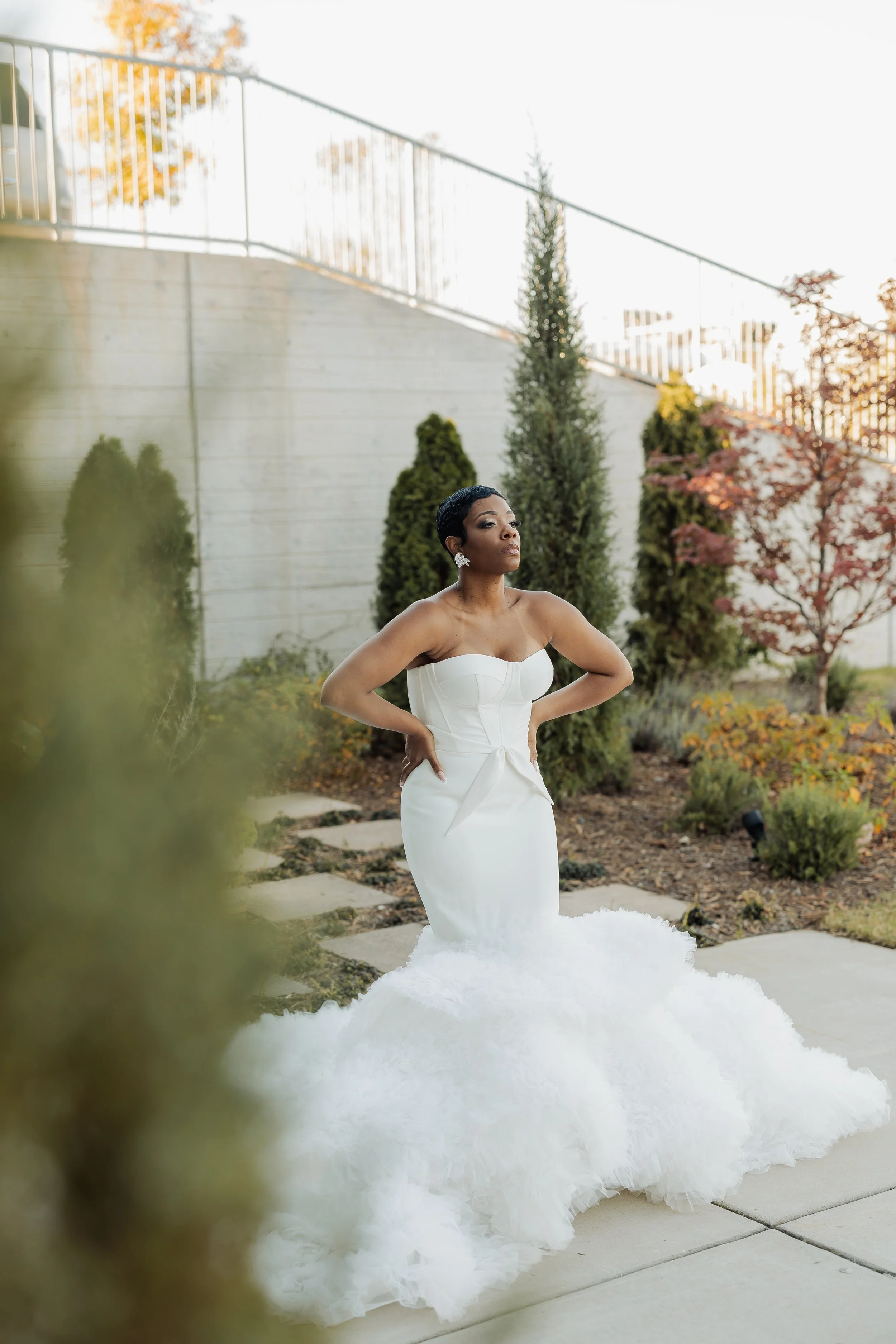 A woman in a strapless white wedding dress with a peplum detail and a long, fluffy train stands outdoors on a concrete pathway, surrounded by trees and bushes with autumn foliage. She has short, curly black hair and is wearing statement earrings, loo