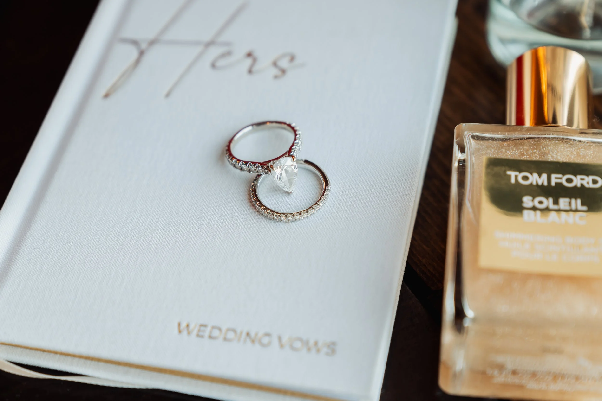 Close-up of a white wedding vow book with gold edges, featuring two rings on top, one with a large gemstone and the other a band with small gemstones, beside a bottle of Tom Ford Soleil Blanc perfume.