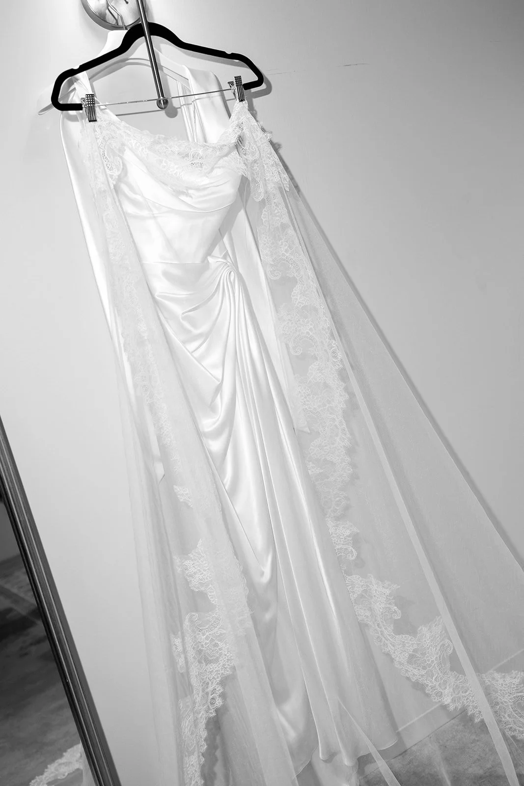 A white wedding dress hanging on a black hanger with a veil attached, displayed on a wall