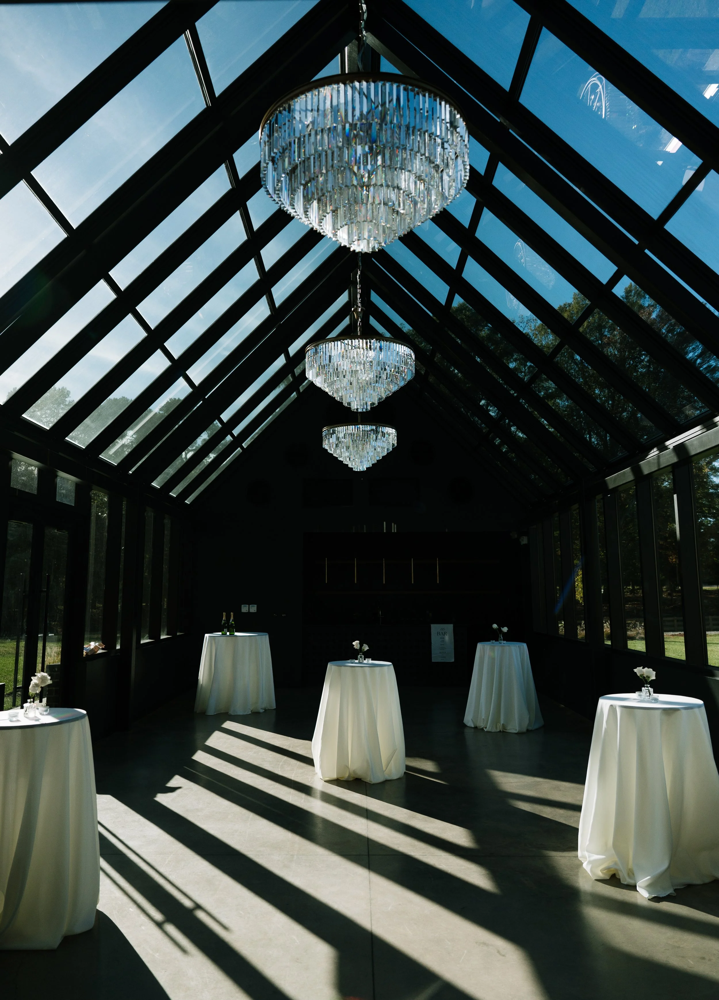 Elegant event space with tall windows, four chandeliers, and high-top tables with white tablecloths and floral centerpieces.