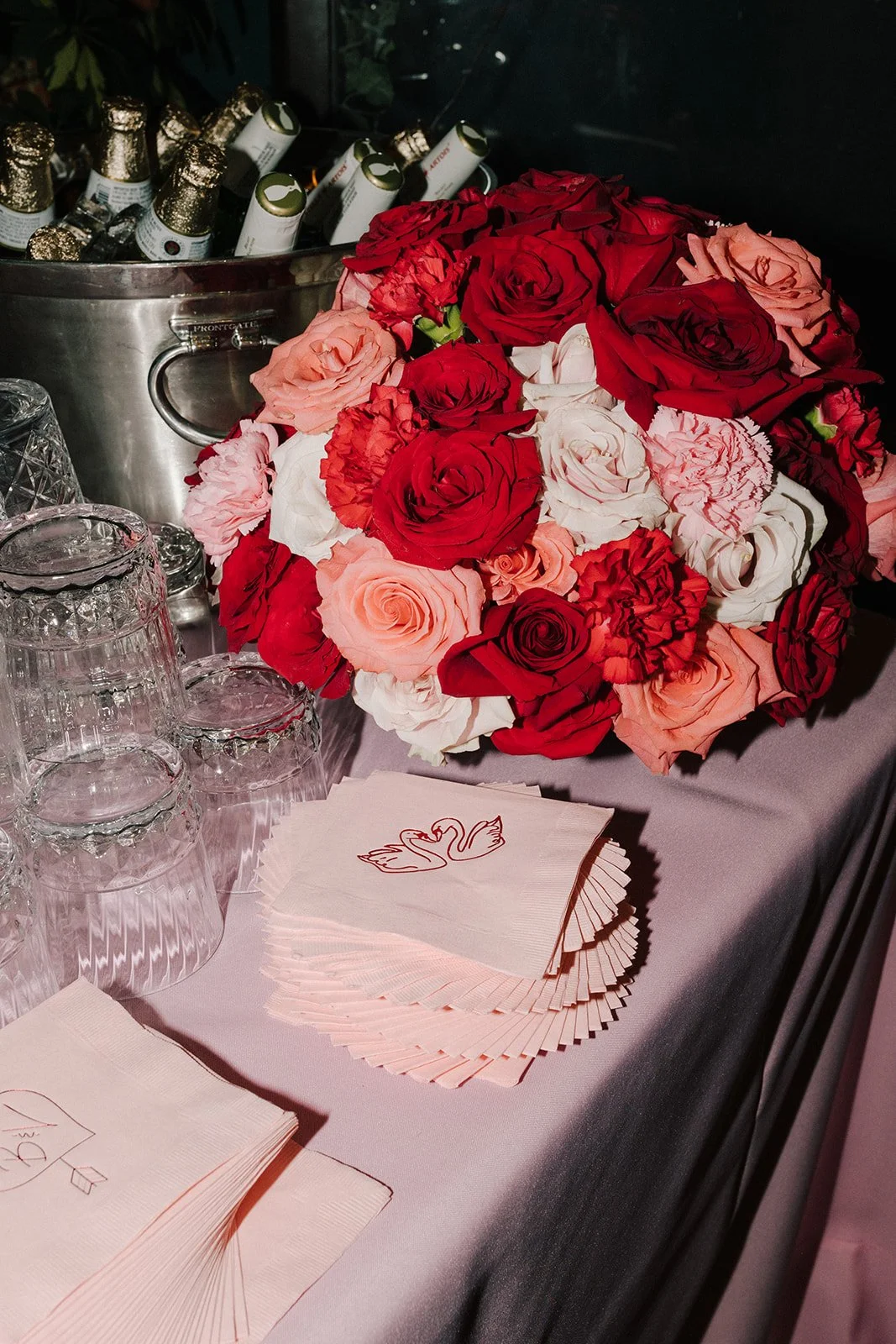 A bouquet of red, pink, and white roses on a table with glassware and swan-themed paper napkins.