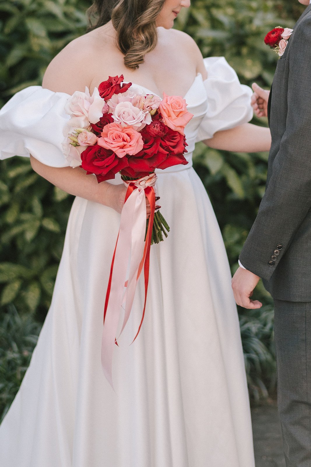 A bride holding a bouquet of red, pink, and white roses during a wedding ceremony, with a groom standing nearby.