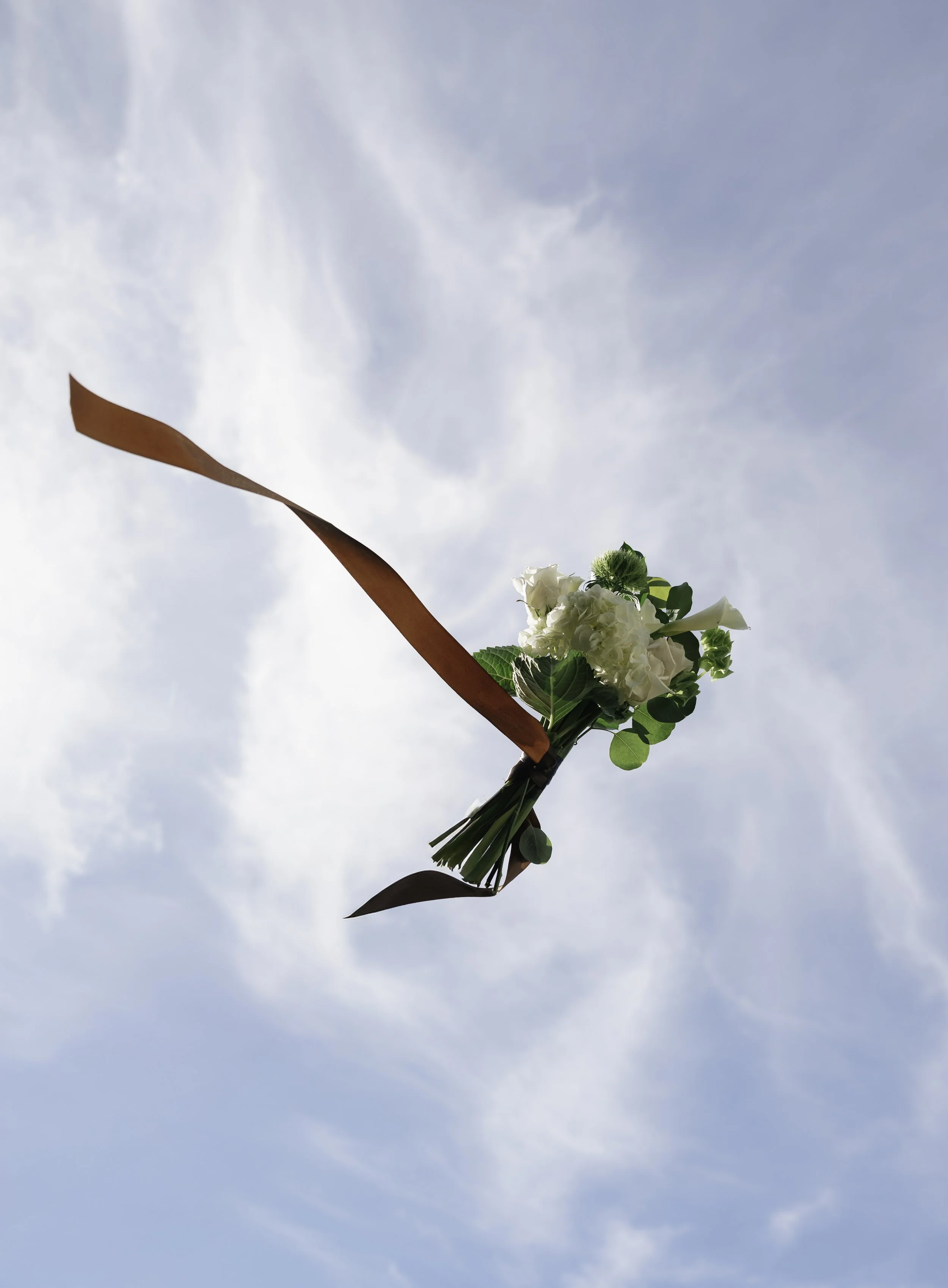 A bouquet of white flowers with green leaves being held up in front of a blue sky with some clouds.