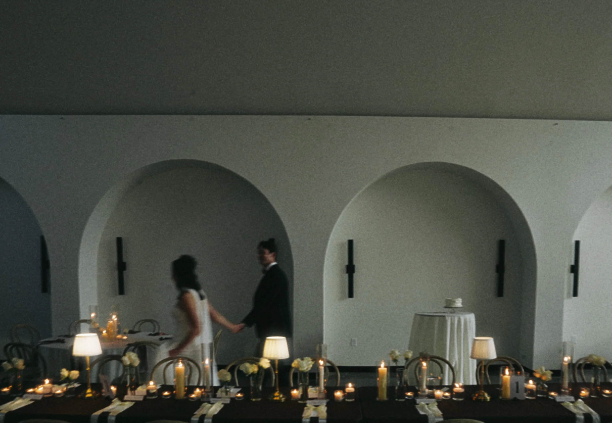 An elegant dining room with tables decorated with candles and flowers, lit by soft lighting, and a white textured ceiling with arched architectural features.