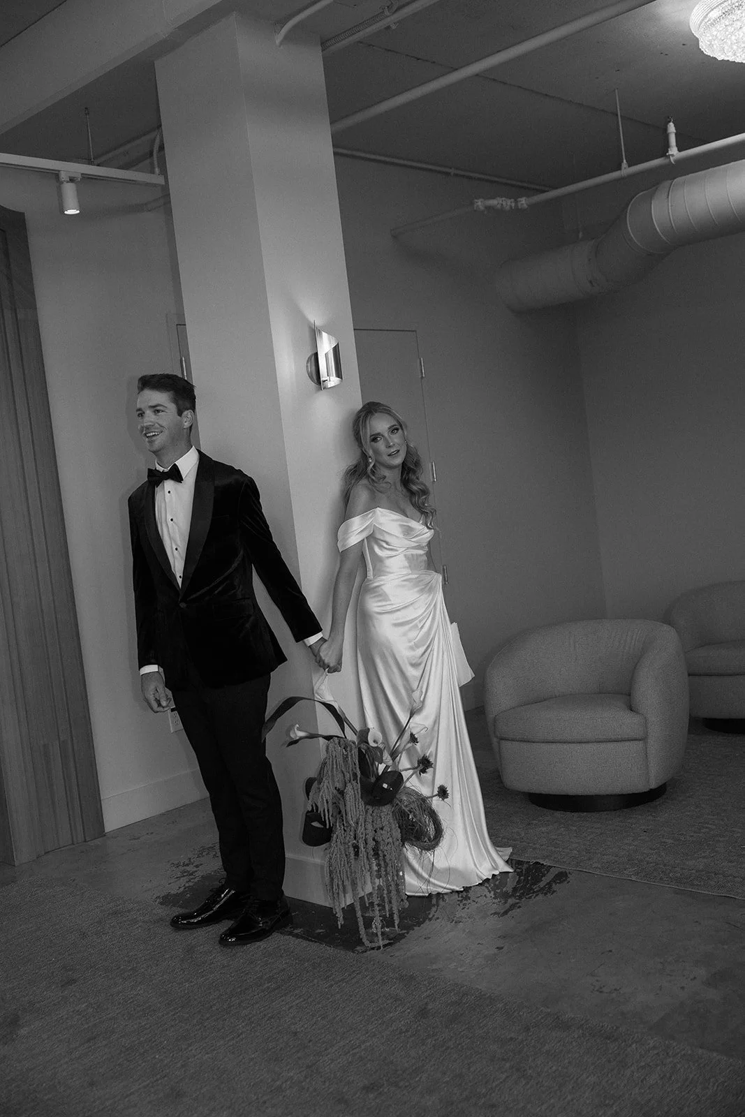 A black and white photo of a man and woman holding hands, with the man dressed in a tuxedo and the woman in a wedding dress, standing in a modern room with furniture and decor.