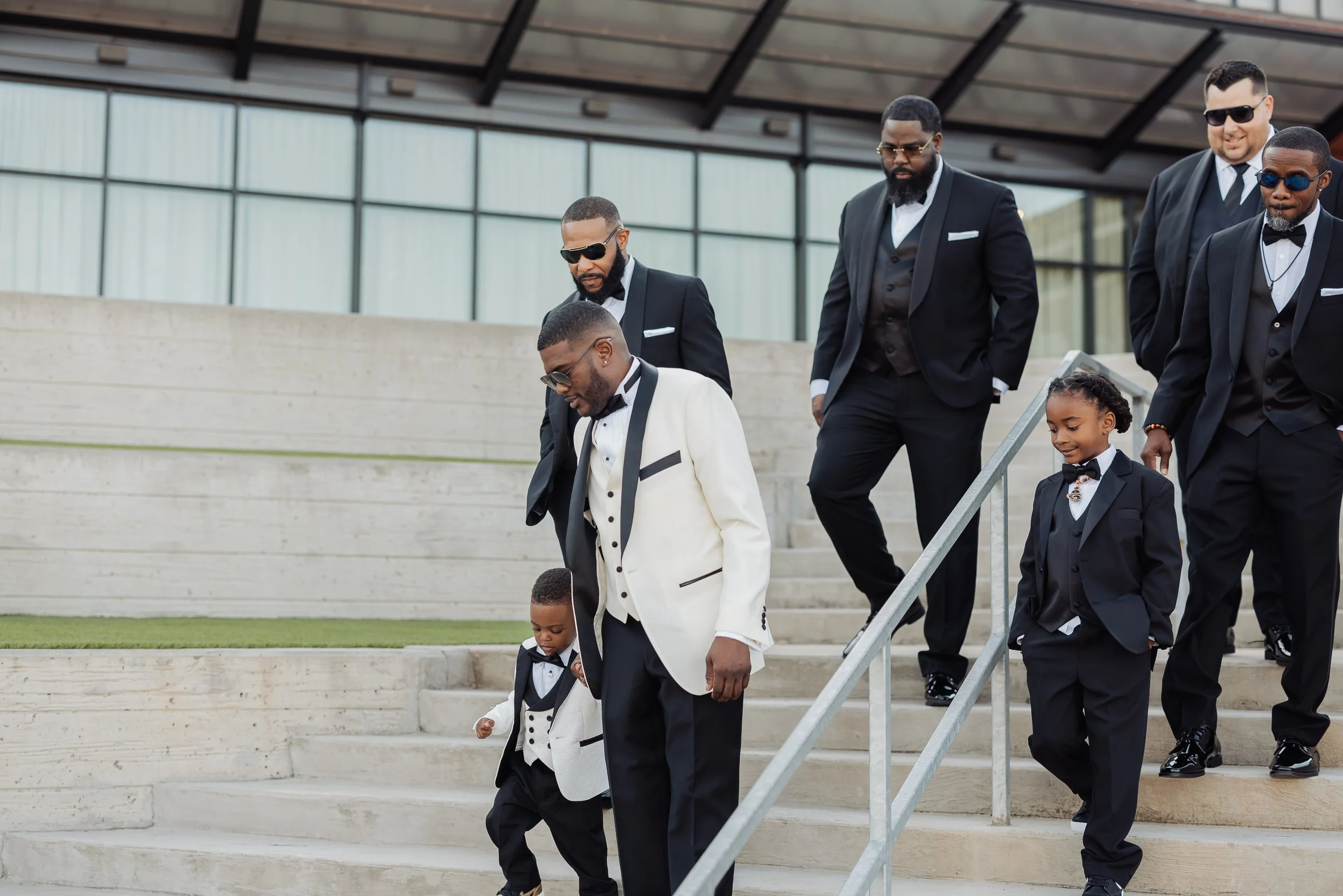 A group of men and a young girl dressed in formal tuxedos and suits, walking down outdoor stairs, possibly attending a wedding or formal event.