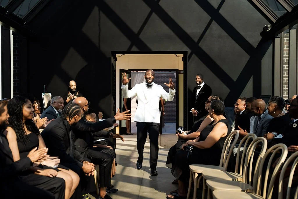 A man in a white tuxedo walks down the aisle at a wedding ceremony, smiling and greeting guests who are seated on either side in a modern, geometric-lit space with black walls.