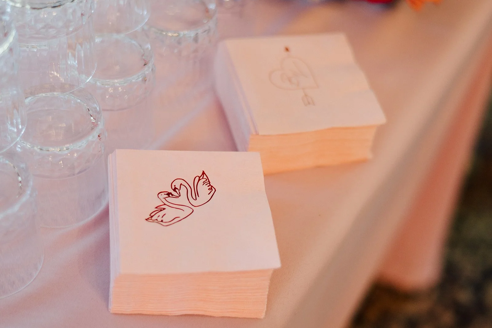 Close-up of a table with vertically stacked pink paper napkins, topped with a white napkin featuring a red outline drawing of a swan, and a pile of similar napkins in the background. To the left, several stemmed glasses are visible.