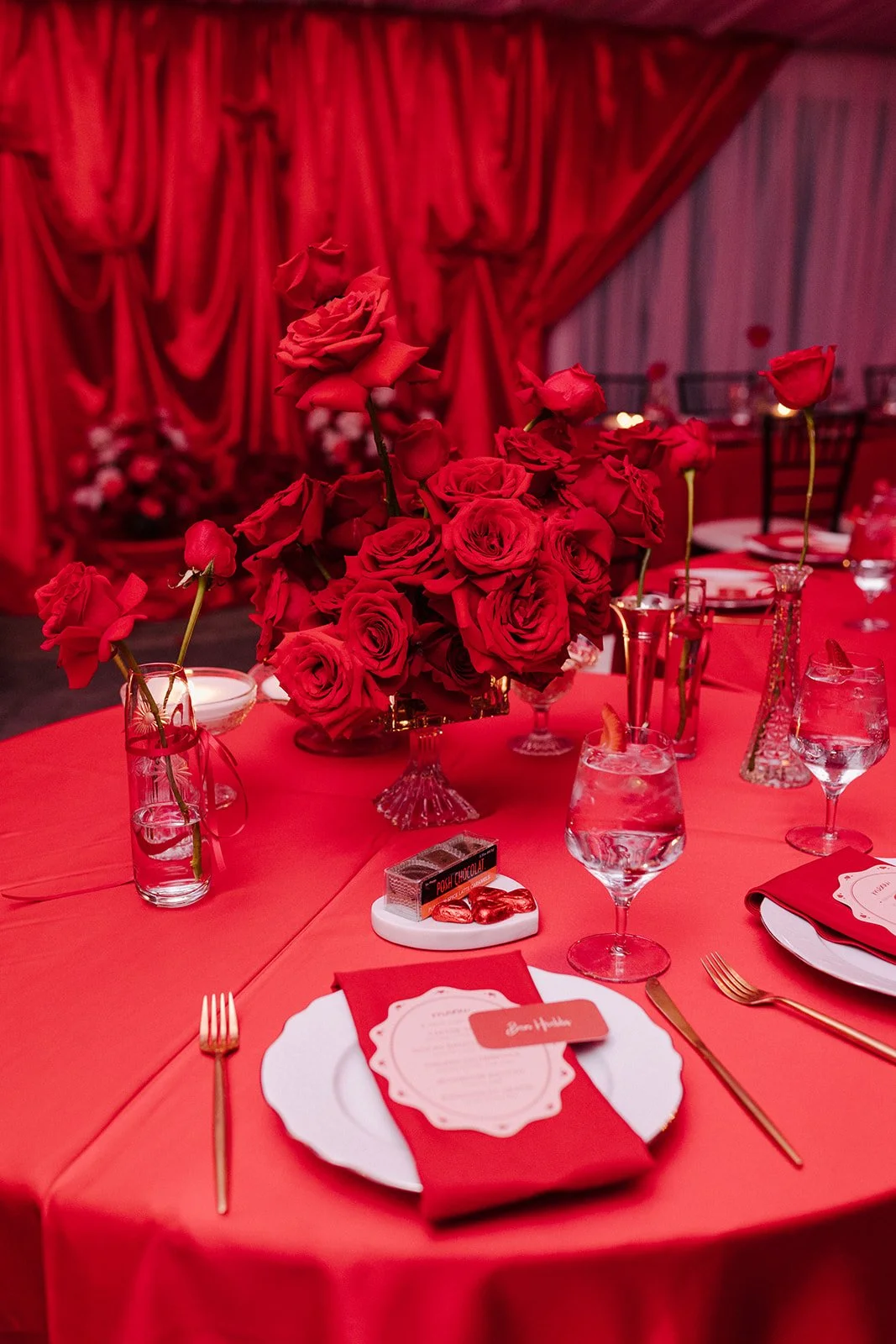 A dining table decorated in shades of red with a centerpiece of red roses in a glass vase, surrounded by glasses, plates, gold flatware, and red napkins. The background features red drapes and a wall with red and pink accents, creating a romantic set